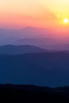 Na obrazie widzimy spokojny krajobraz górski o wschodzie lub zachodzie słońca. Słońce znajduje się po prawej stronie, nisko na horyzoncie, oświetlając niebo delikatnymi odcieniami różu, pomarańczu i fioletu. Góry w oddali układają się w łagodne, stopniowane warstwy, każda kolejna ciemniejsza, co tworzy głębię i atmosferę spokoju.