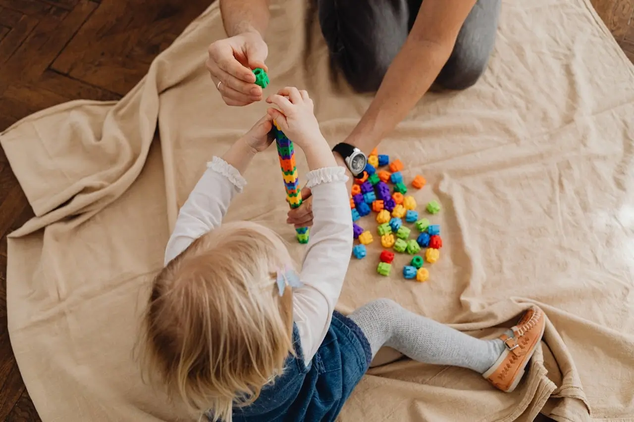 Małe dziecko bawiące się kolorowymi klockami edukacyjnymi z dorosłym na jasnym kocu, widok z góry, scena wspólnej zabawy i nauki.