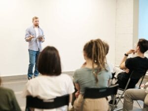 Mężczyzna na scenie w czasie wykładu i fragment siedzącej publiczności.